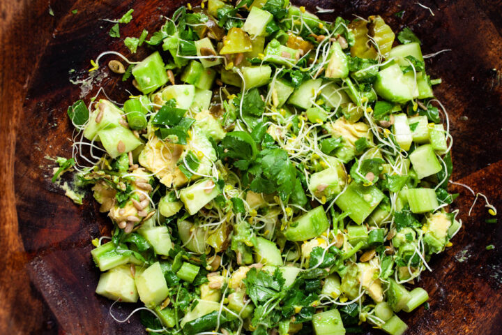 A green salad with sweet pickles, cucumber, green onions, avocado, pumpkin seeds, cilantro leaves, and broccoli sprouts seasoned and served in a wooden bowl.