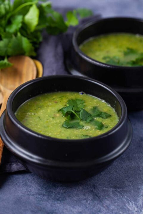 Two black bowls filled with green pea soup, topped with fresh Parsley leaves next to two wooden spoons and more fresh Parsley.