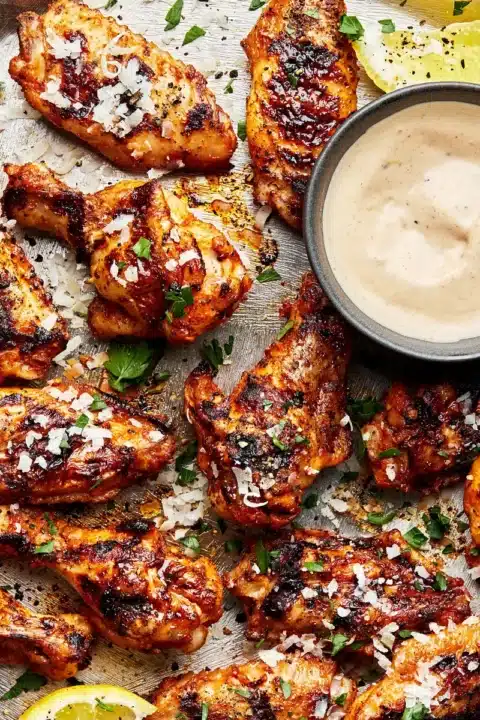 an overhead shot of grilled lemon pepper chicken whites on a baking tray beside a bowl of sauce and lemon wedges tucked in amongst the wings.