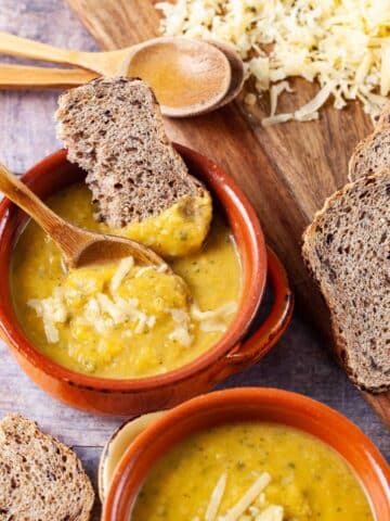 Two bowls filled with yellow squash soup, topped with Parmesan cheese and served with extra cheese and bread on the side.