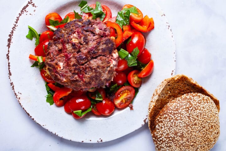 A cooked hamburger served on top of a healthy salad and a bun on the side.