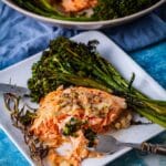 A fork resting on a plate with a piece of baked salmon and a bit of baby broccoli, arranged next to a slab of the pierced in baked salmon and baked broccoli with a sprig of baked thyme on the side.