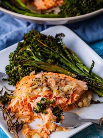 A fork resting on a plate with a piece of baked salmon and a bit of baby broccoli, arranged next to a slab of the pierced in baked salmon and baked broccoli with a sprig of baked thyme on the side.