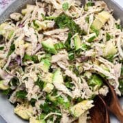 Two wooden spoons resting in a large plate filled with a healthy chicken salad with avocado.