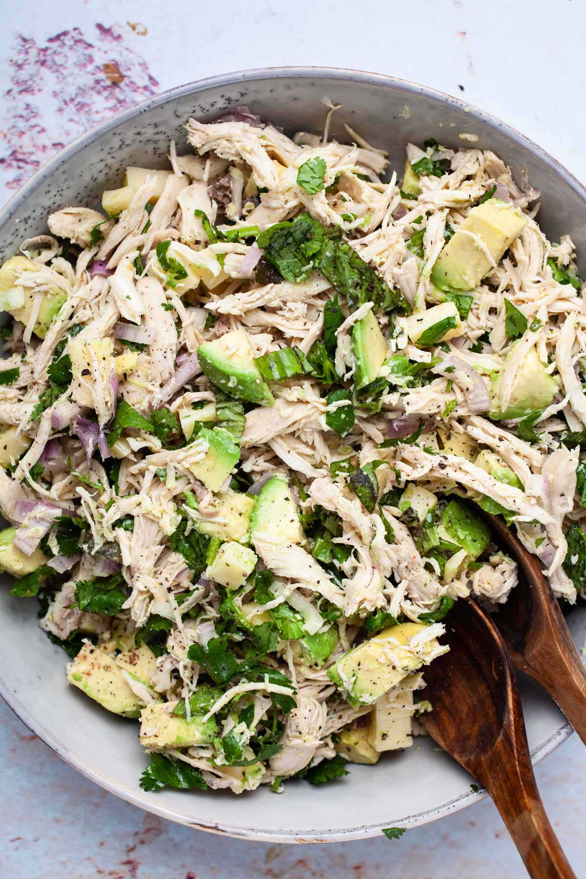 Two wooden spoons resting in a large plate filled with a healthy chicken salad with avocado.