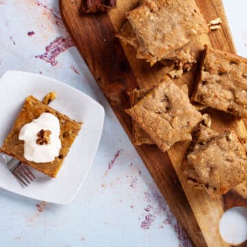 Squares of coffee cake stacked on a wooden board next to one on a plate topped with creamy yogurt.