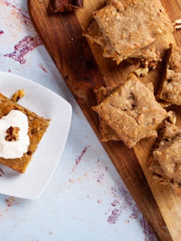 Squares of coffee cake stacked on a wooden board next to one on a plate topped with creamy yogurt.