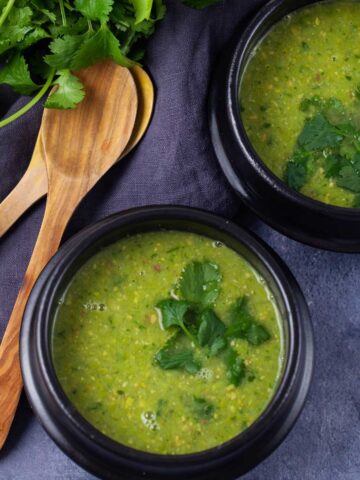 Two black bowls filled with green pea soup, topped with fresh Parsley leaves next to two wooden spoons and more fresh Parsley.