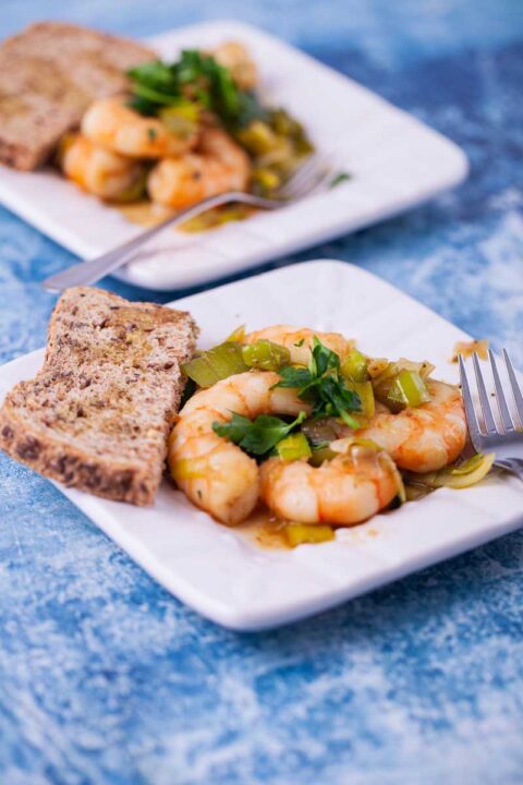 A close up of sauteed shrimp with leeks on a small plate served with toast and a drizzle of olive oil.