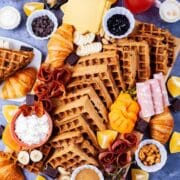 Fresh fruits, cheeses, charcuterie, meats, chocolate, croissants, spreads, coffee, juices, nuts, and homemade waffles all beautifully displayed on a waffle board and table.