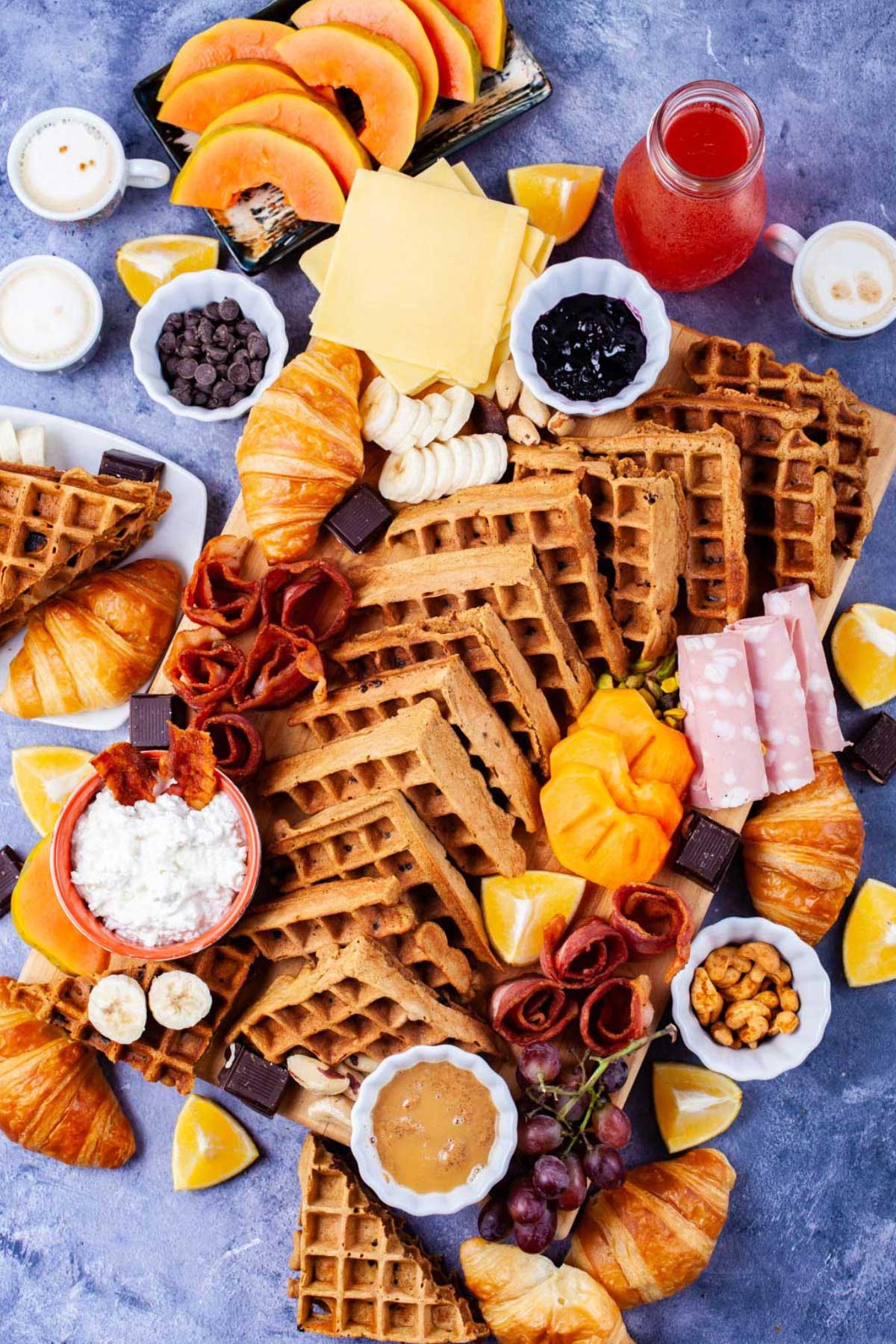 Fresh fruits, cheeses, charcuterie, meats, chocolate, croissants, spreads, coffee, juices, nuts, and homemade waffles all beautifully displayed on a waffle board and table.