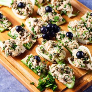 Fresh cucumber and fennel slices topped with tuna salad and garnished with dark grapes neatly presented on a wooden board.