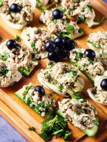 Fresh cucumber and fennel slices topped with tuna salad and garnished with dark grapes neatly presented on a wooden board.