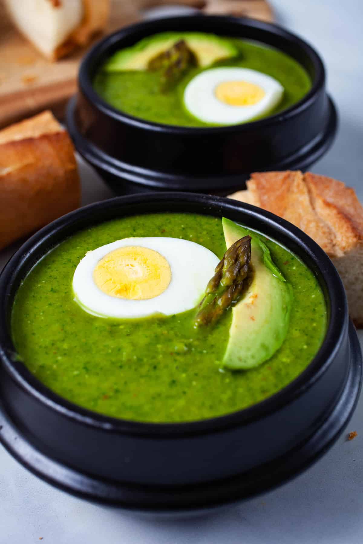 Two bowls of asparagus soup topped with avocado, half of a cooked egg and head of an asparagus.