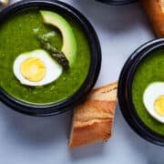 Two bowls of asparagus soup topped with avocado, half of a cooked egg and head of an asparagus.
