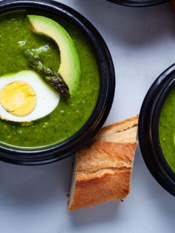 Two bowls of asparagus soup topped with avocado, half of a cooked egg and head of an asparagus.