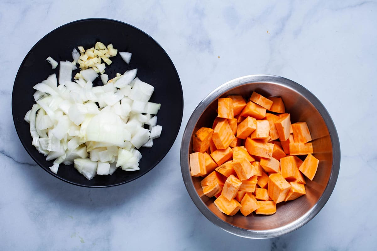 Chopped sweet potatoesin a bowl next to chopped onions and garlic in a plate.