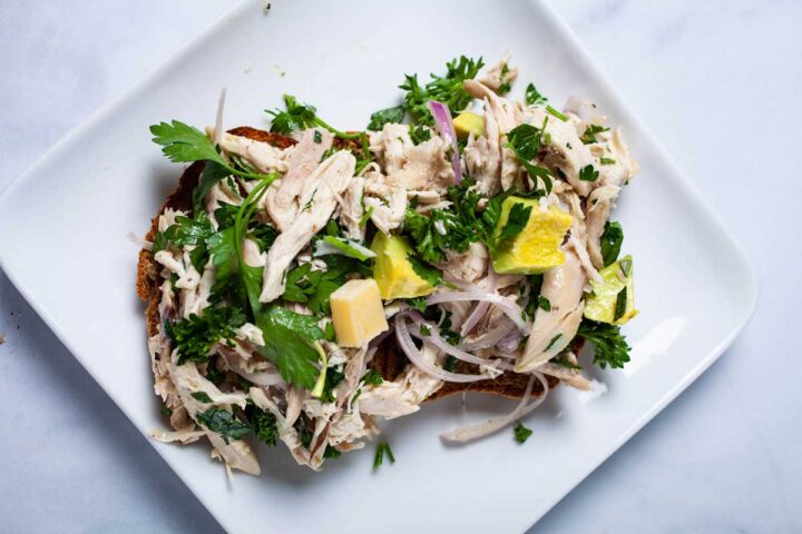 A small plate with Keto bread topped with healthy chicken salad infused with fresh chopped cilantro, parsley, avocado, hard cheese, and sliced shallots, and seasoned with olive oil, salt, and pepper.