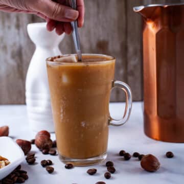 A hand stirring a spoon in a tall glass coffee cup filled with a cold iced latte.