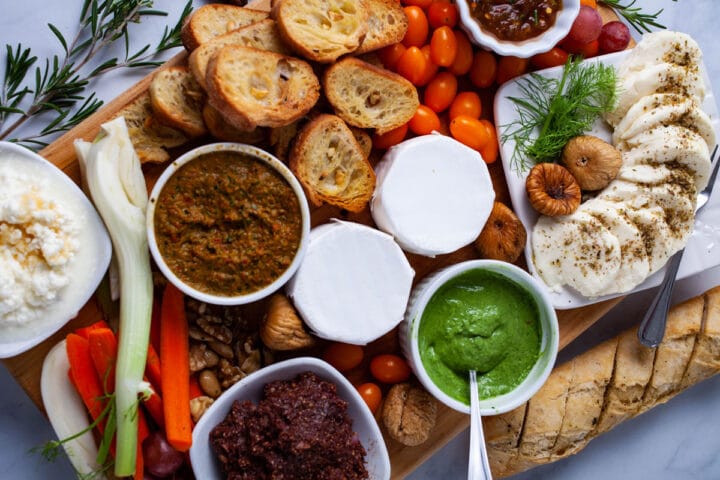 A wooden board topped with Italian dips, freshly cut veggies, fruits, cheeses, dried fruits, crostini bread, and spreads.