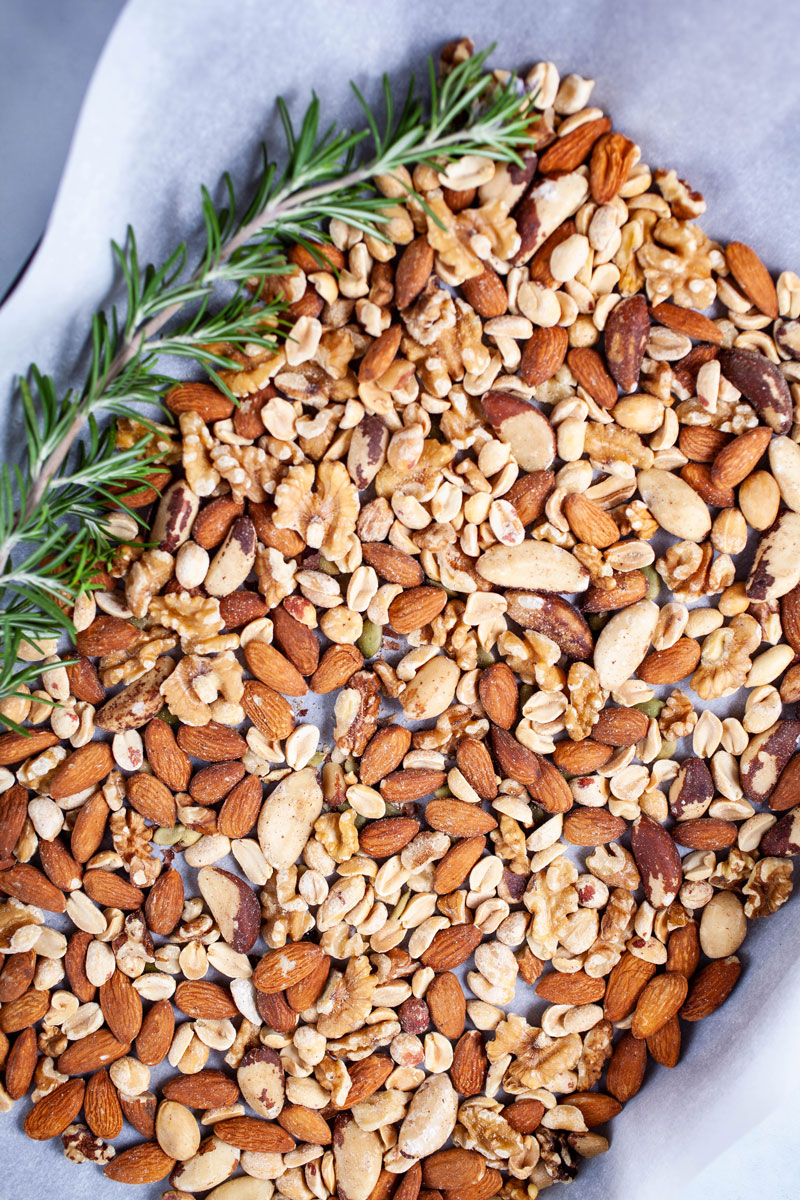 A mix of nuts and a large Rosemary leave spread out on a prepared baking sheet.