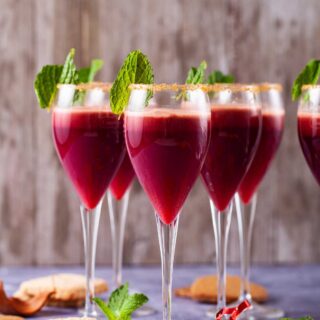 A close-up of champagne glasses with a sugary rim, filled with a red drink, garnished with mint leaves and served with cookies on the side.