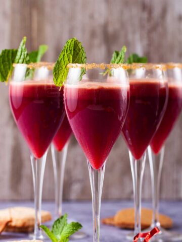 A close-up of champagne glasses with a sugary rim, filled with a red drink, garnished with mint leaves and served with cookies on the side.