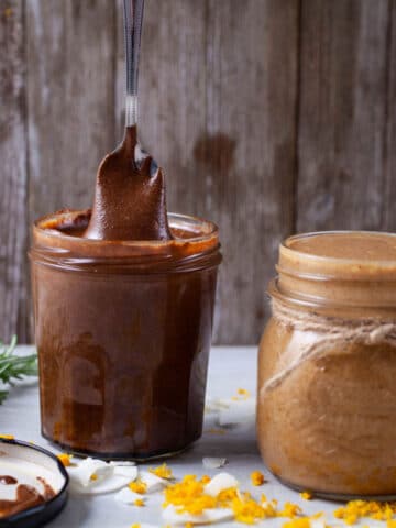 A small spoon lifting creamy chocolate nut butter out of a jar and next to another jar with coconut butter.