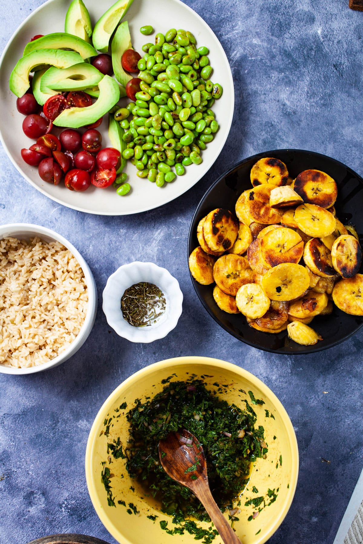 Cooked rice, tomatoes, sliced avocado, fried edamame beans, fried plantains, chimichurri sauce, and seasoning displayed on a table.