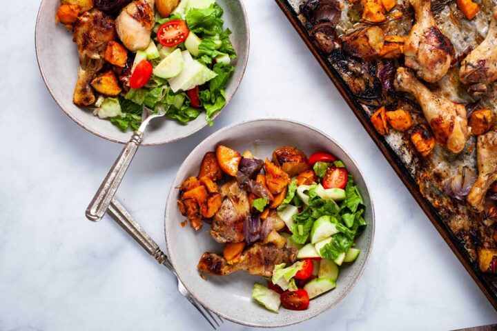 Two plates filled with roasted drumsticks, sweet potatoes and a green salad next to a chicken dinner sheet pan.