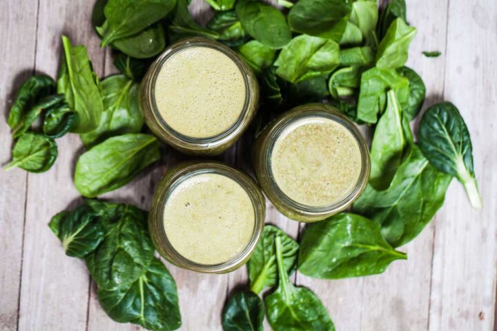 Three small glass jars filled with green smoothie and surrounded by fresh spinach leaves.