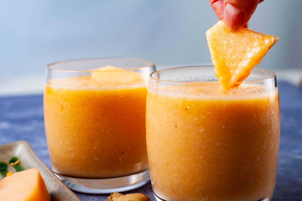 A hand holding a piece of melon above a cantaloupe smoothie to dip into.
