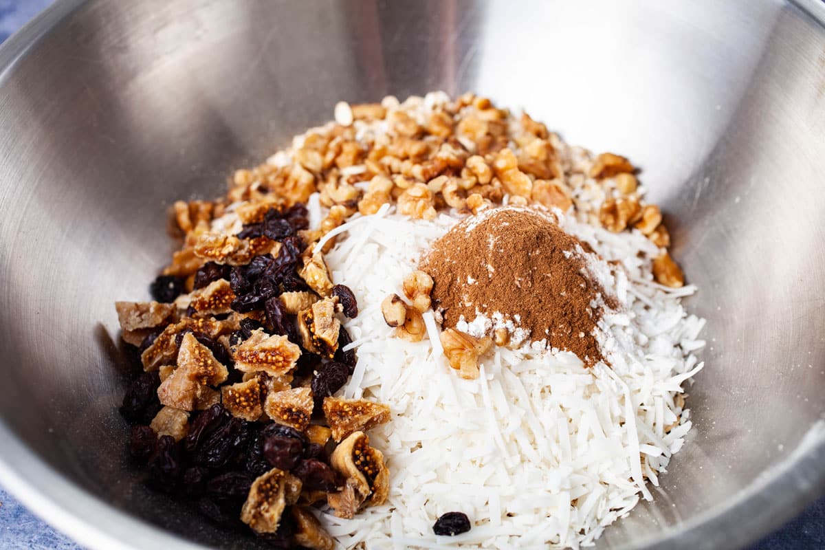 A stainless steel bowl with oats, chopped walnuts, raisins, chopped figs, coconut flakes, and cinnamon powder.