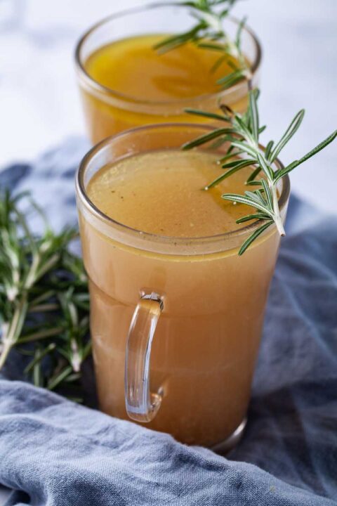 Two cups filled with broth and topped with fresh Rosemary leaves.