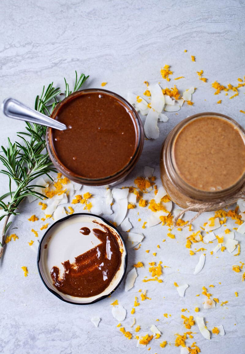 Creamy chocolate nut butter and coconut butter in mason jars and surrounded by Rosemary leaves, coconut shreds, and orange zest.