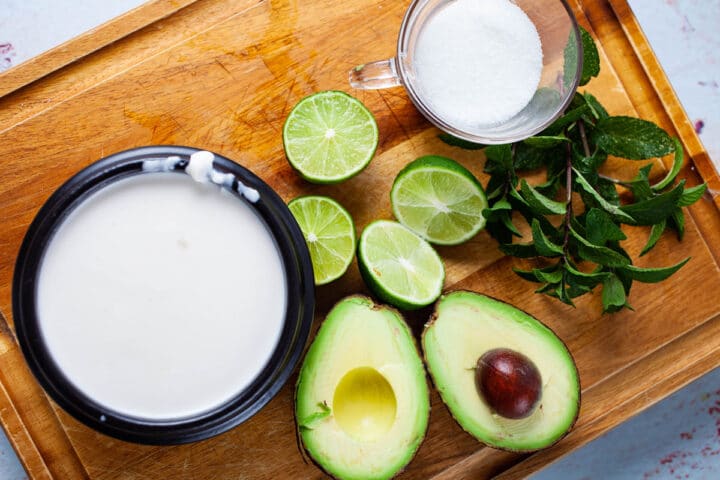 Coconut cream, one avocado cut in half, two limes cut in half, Keto sugar, and fresh mint leaves displayed on a wooden board to make healthy popsicles.