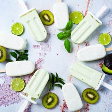 Green and white popsicles displayed in a circle on a table and decorated with slices of kiwi, lime, avocado, and mint leaves.