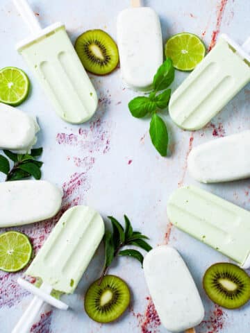 Green and white popsicles displayed in a circle on a table and decorated with slices of kiwi, lime, avocado, and mint leaves.