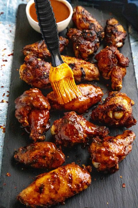 a grilling brush brushing on a honey sriracha sauce on chicken wings that are arranged on a black serving board.
