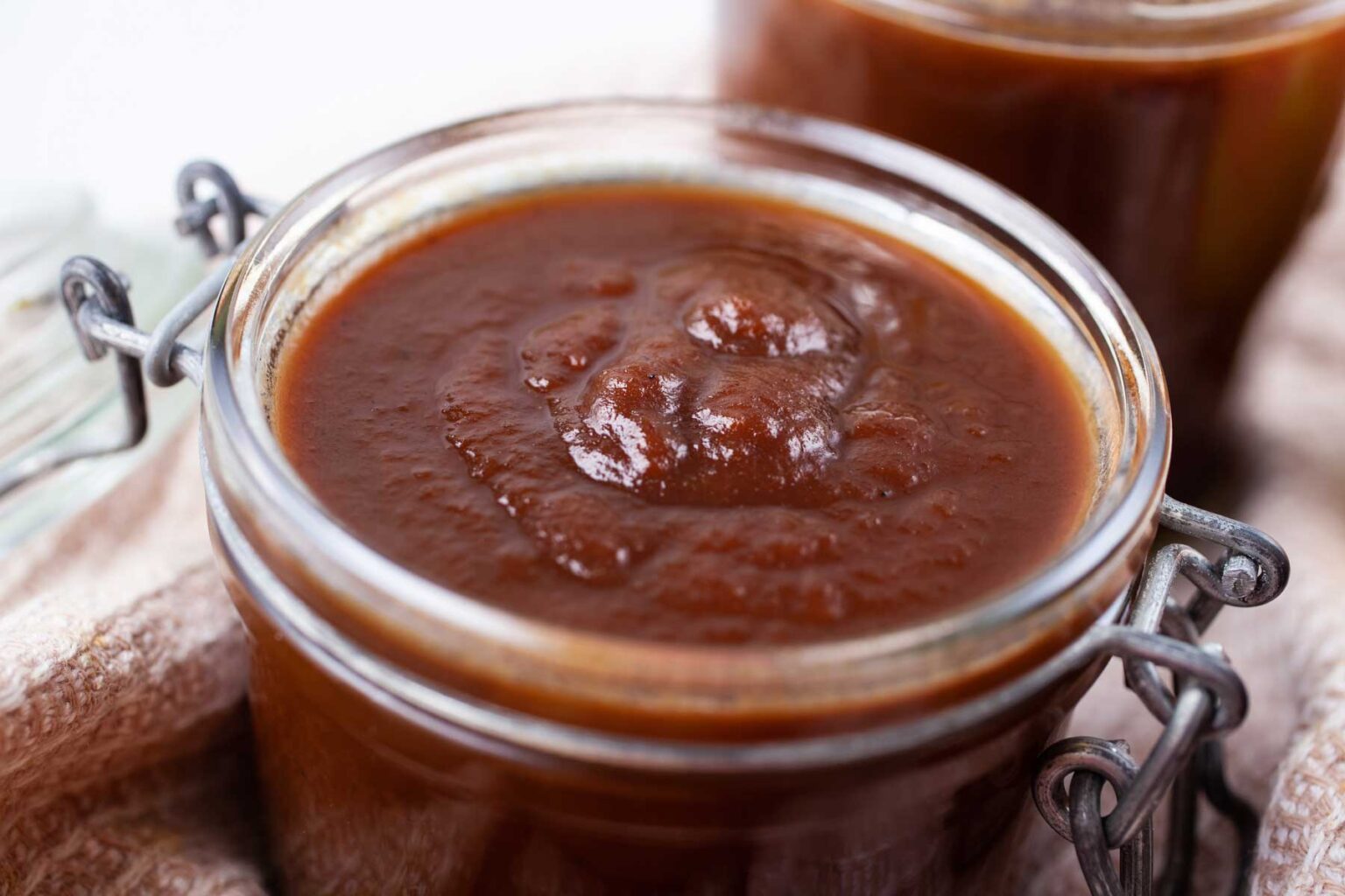 A close-up of a glass jar filled with thick, reddish-brown sauce, likely barbecue sauce or ketchup. The jar has a metal clasp lid and sits on a beige cloth with another jar in the blurred background.