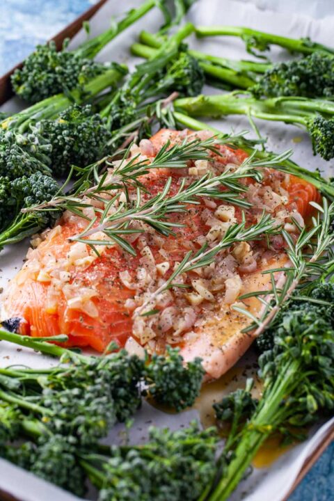 Raw salmon fillet, surrounded by baby broccoli, and topped with onions and butter arranged on a baking sheet.