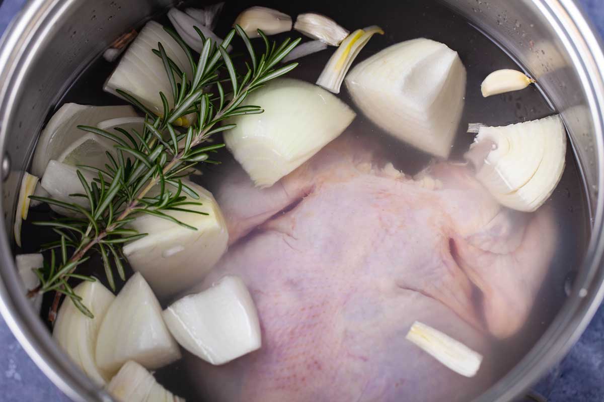 A large pot filled with a raw chicken, chopped onions, garlic, and fresh Rosemary leaves.