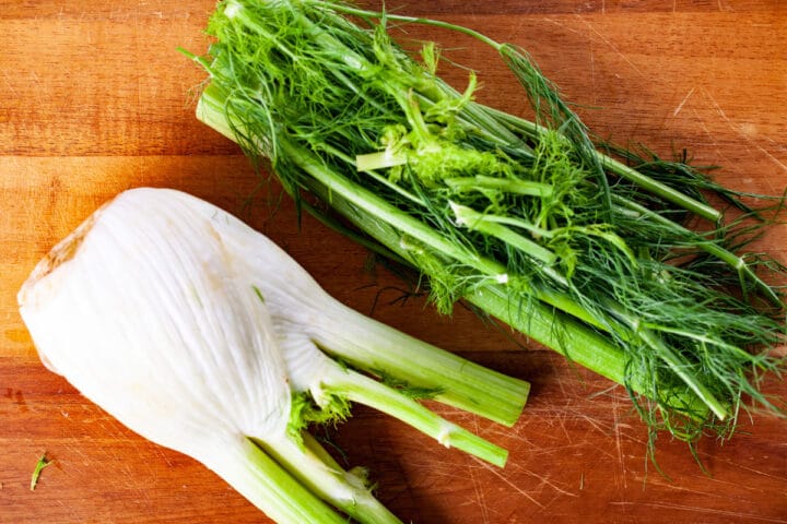 A raw fennel bulb on a cutting board with its fennel fronds removed and displayed next to it.