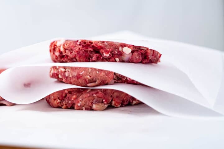 Three raw prepped hamburgers stacked on top of each other and separated with parchment paper.