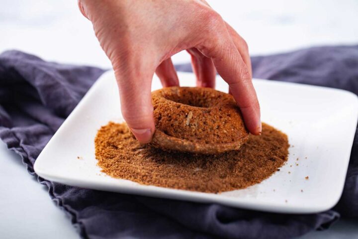 A hand dipping a baked donut into a cinnamon sugar mix.