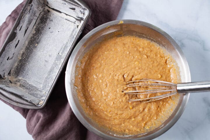 A whisk resting in a bowl with cake batter next to a cake loaf pan.