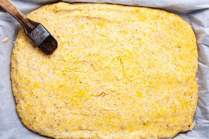 A kitchen brush resting on a polenta pizza crust coated with olive oil.