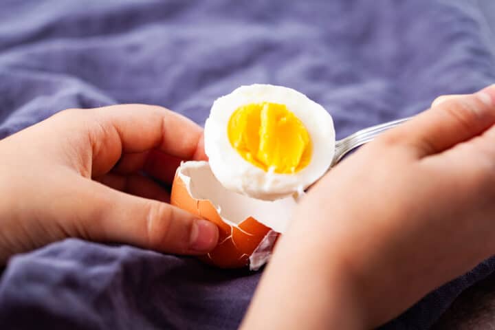 A child's hands lifting half of a cooked egg out of its shell with a spoon.