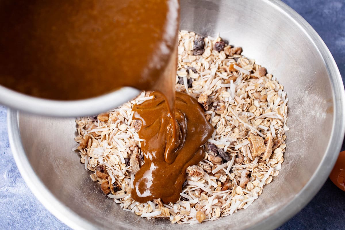 A bowl dripping a brown sweet paste into a stainless steel bowl containing an oat mixture.