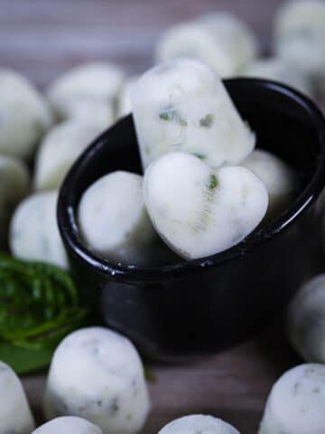 A close up of white ice cubes in a small bowl and surrounded by randomly placed ice cubes on a wooden board.